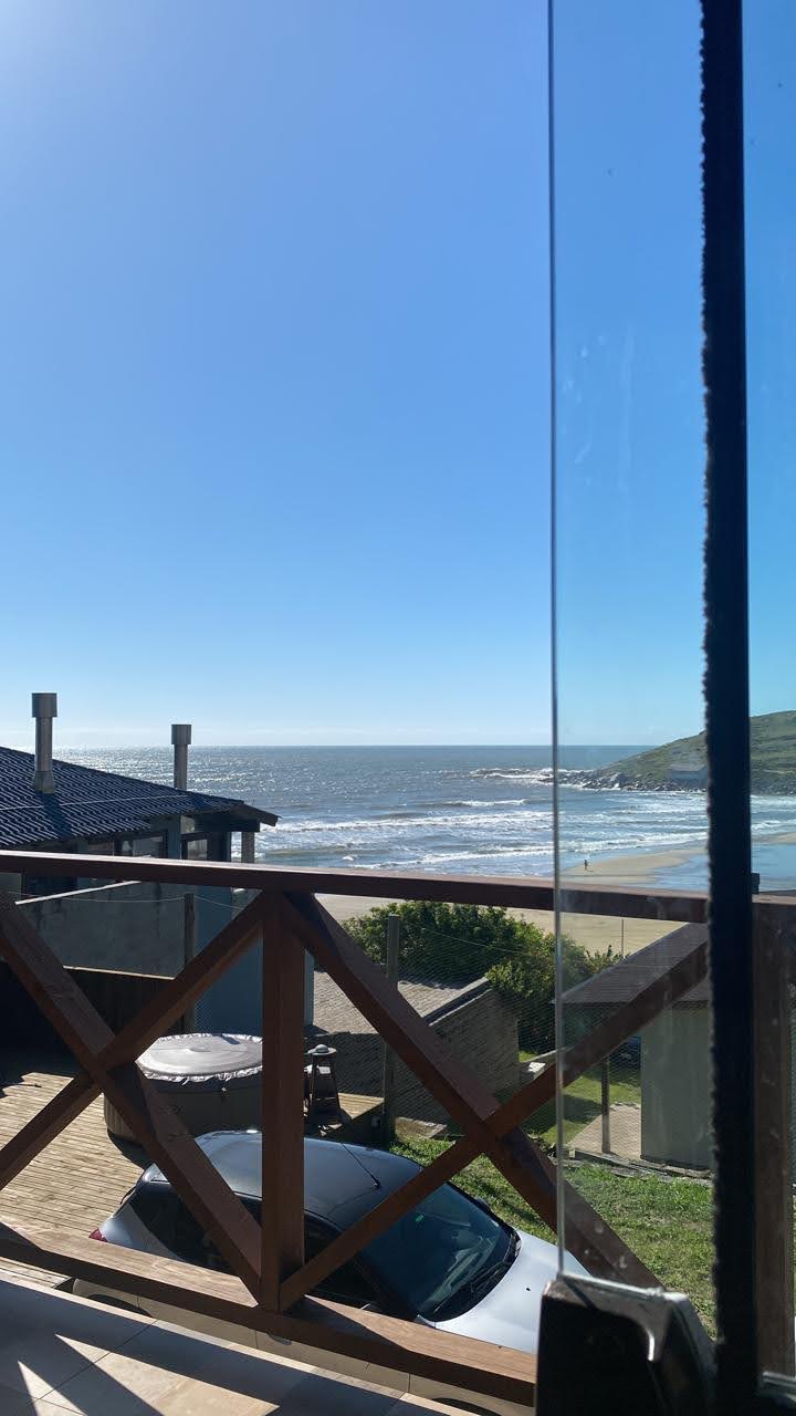 A panoramic view captures the ocean and waves beyond a wooden railing, with a clear sky above. A car is parked on the stone path, and the lush green landscape is visible beside the beach.