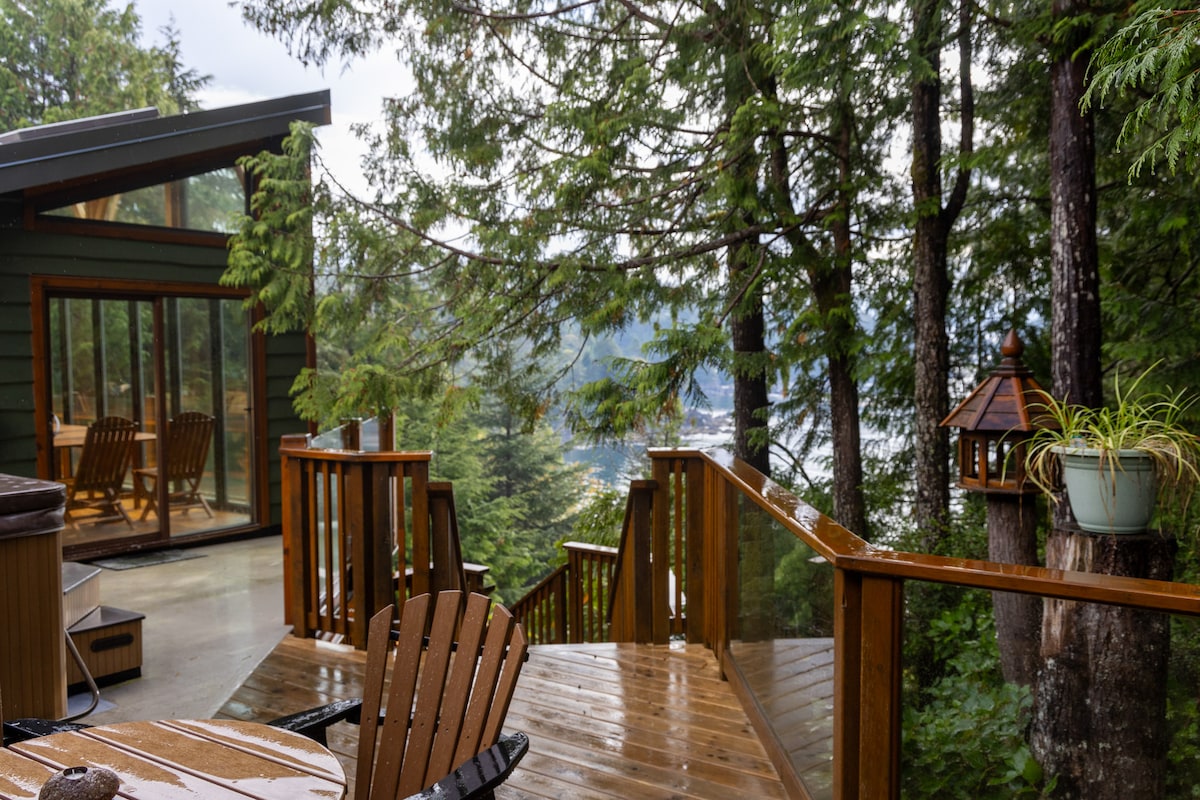 A wooden deck featuring a round table and chairs is surrounded by tall trees, with views of the distant ocean through the greenery. The nearby space showcases a large glass sliding door leading into a cozy building, enhancing the connection with nature.