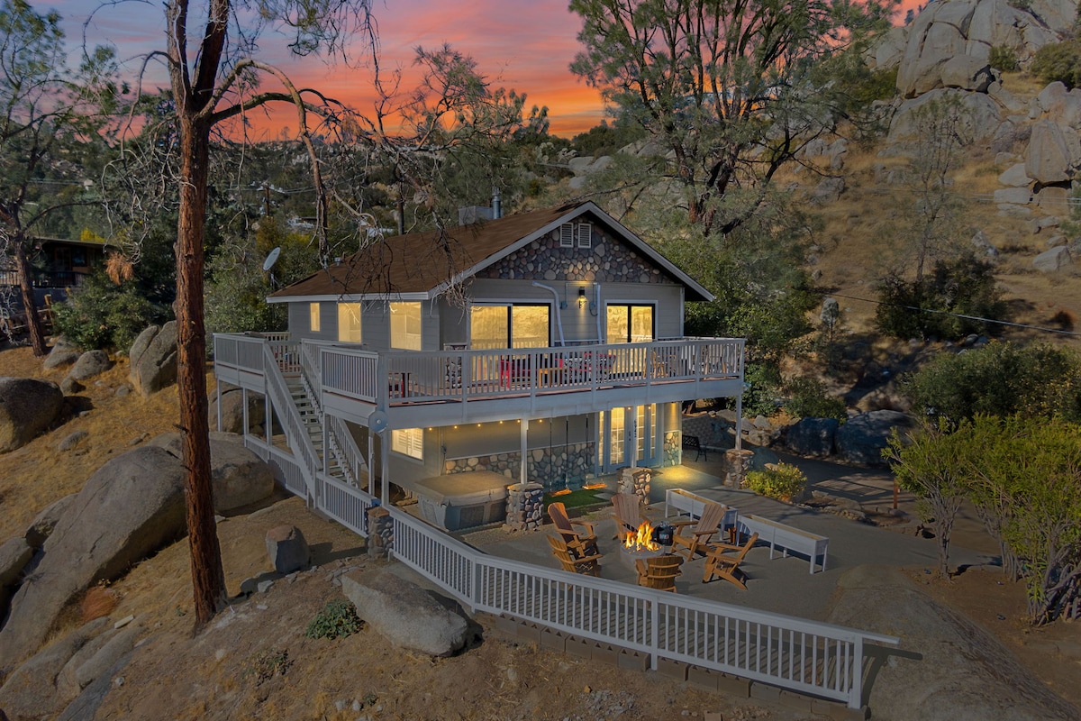 A two-story house is situated among natural scenery, featuring a spacious deck with seating and a firepit. Large windows illuminate the interior, showcasing a warm ambiance at sunset. The surrounding landscape includes boulders and trees, enhancing the tranquil outdoor setting.