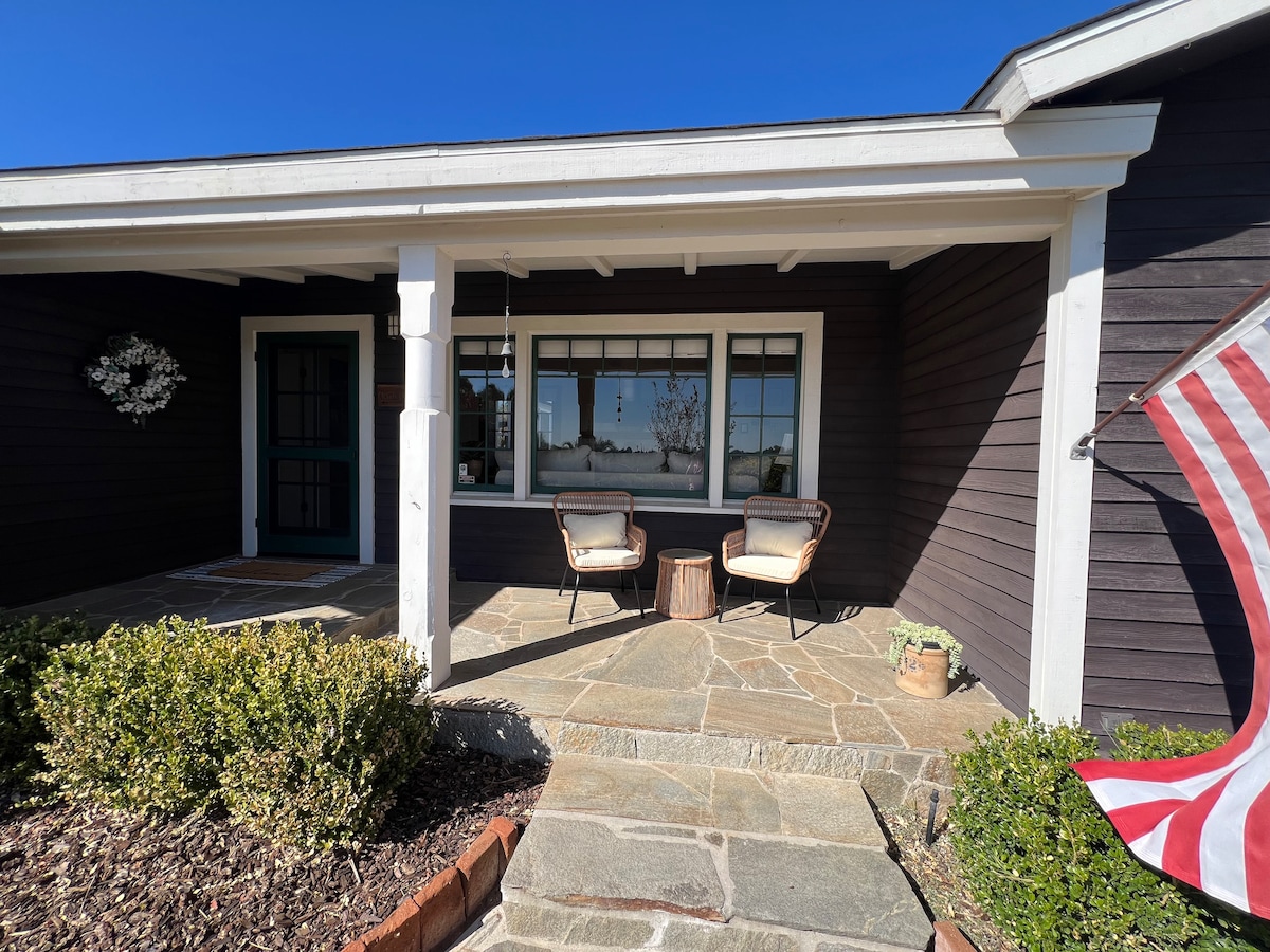 An inviting porch area features two seated chairs and a small wooden side table, set on a stone pathway. A welcoming entrance is framed by white columns and a vibrant blue sky serves as the backdrop, while landscaping includes low-growing green plants.