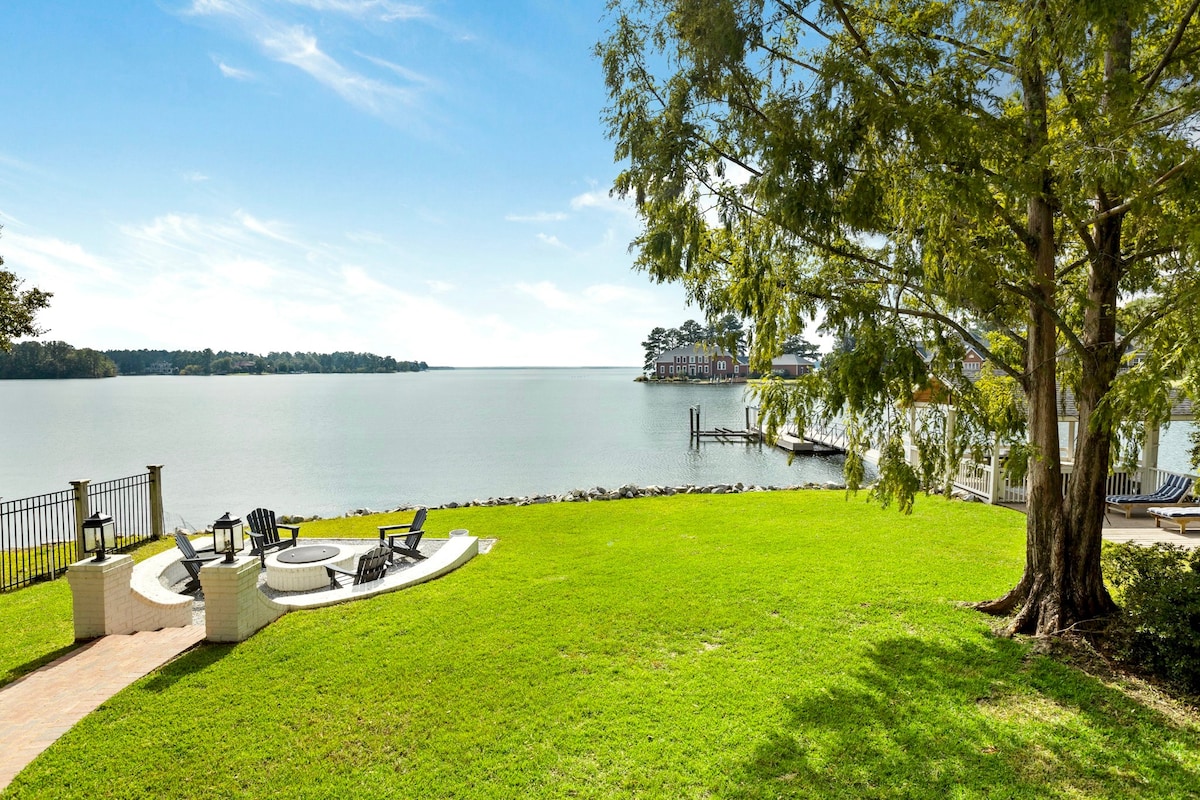 A serene lakeside view showcases a well-maintained lawn leading to the water's edge. A circular seating area surrounding a gas fire pit is positioned on the grassy area, creating an inviting space for relaxation. Trees frame the scene, enhancing the natural setting alongside the lake.