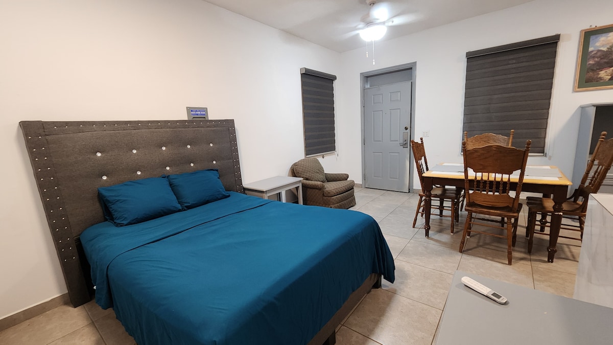 A spacious room features a king bed adorned with blue bedding against a gray upholstered headboard. A seating area includes an armchair, and a dining table with six wooden chairs occupies one corner. Two windows are covered by dark shades.