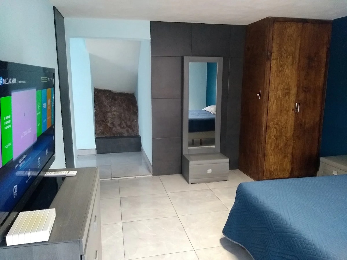 A comfortable bedroom features a spacious bed covered with a blue quilt. A large flat-screen television is mounted on the wall opposite. A mirror and a modest dresser are situated next to the bed, enhancing storage options. A cozy corner is visible in the background.