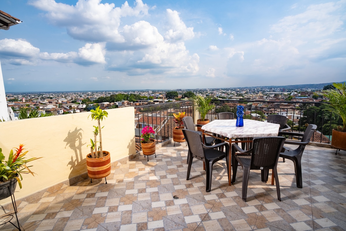 A spacious terrace is featured, furnished with a round dining table surrounded by chairs. Potted plants add a vibrant touch. The panoramic view of the city is visible in the background, enhanced by a bright sky with scattered clouds.