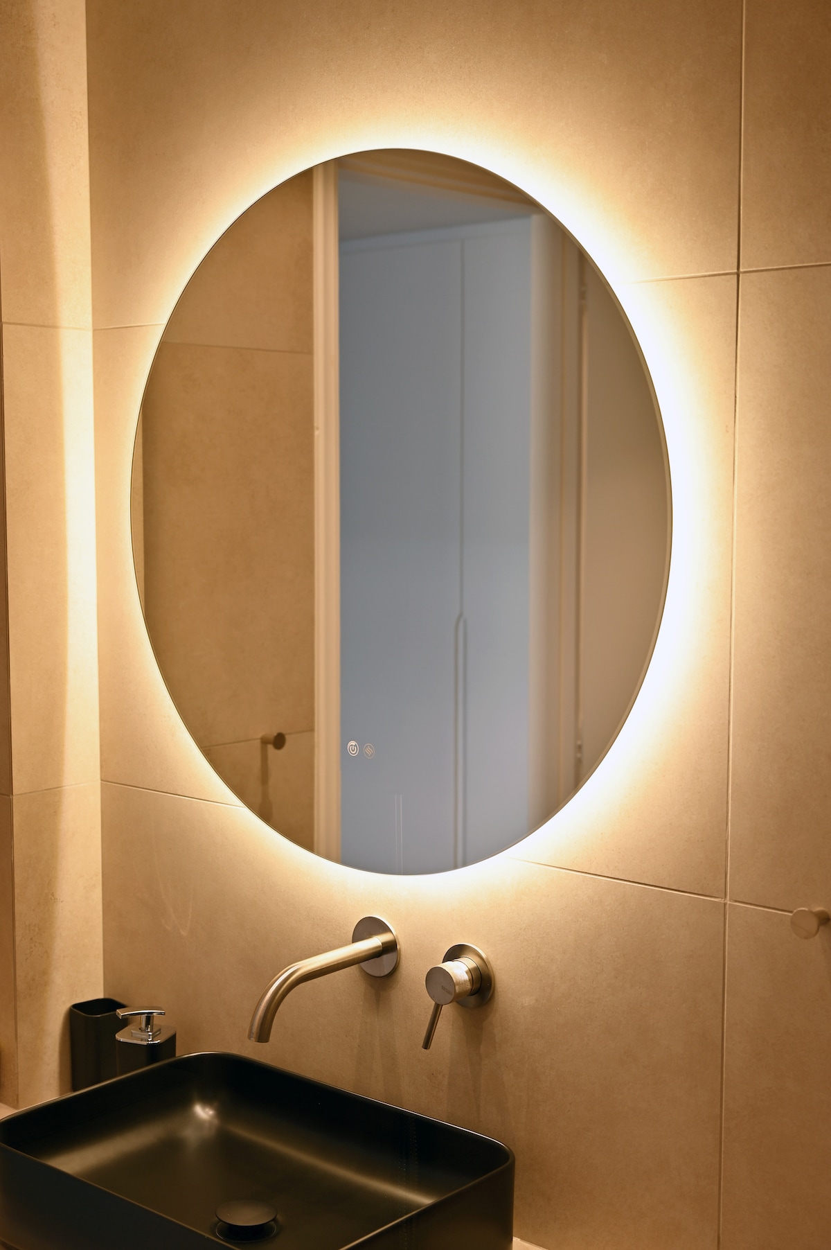 A circular mirror with integrated lighting is displayed above a sleek black sink. The surrounding tiled wall has a warm, neutral tone. The reflection shows a hint of a lighted doorway, providing a modern and functional design element in the bathroom.
