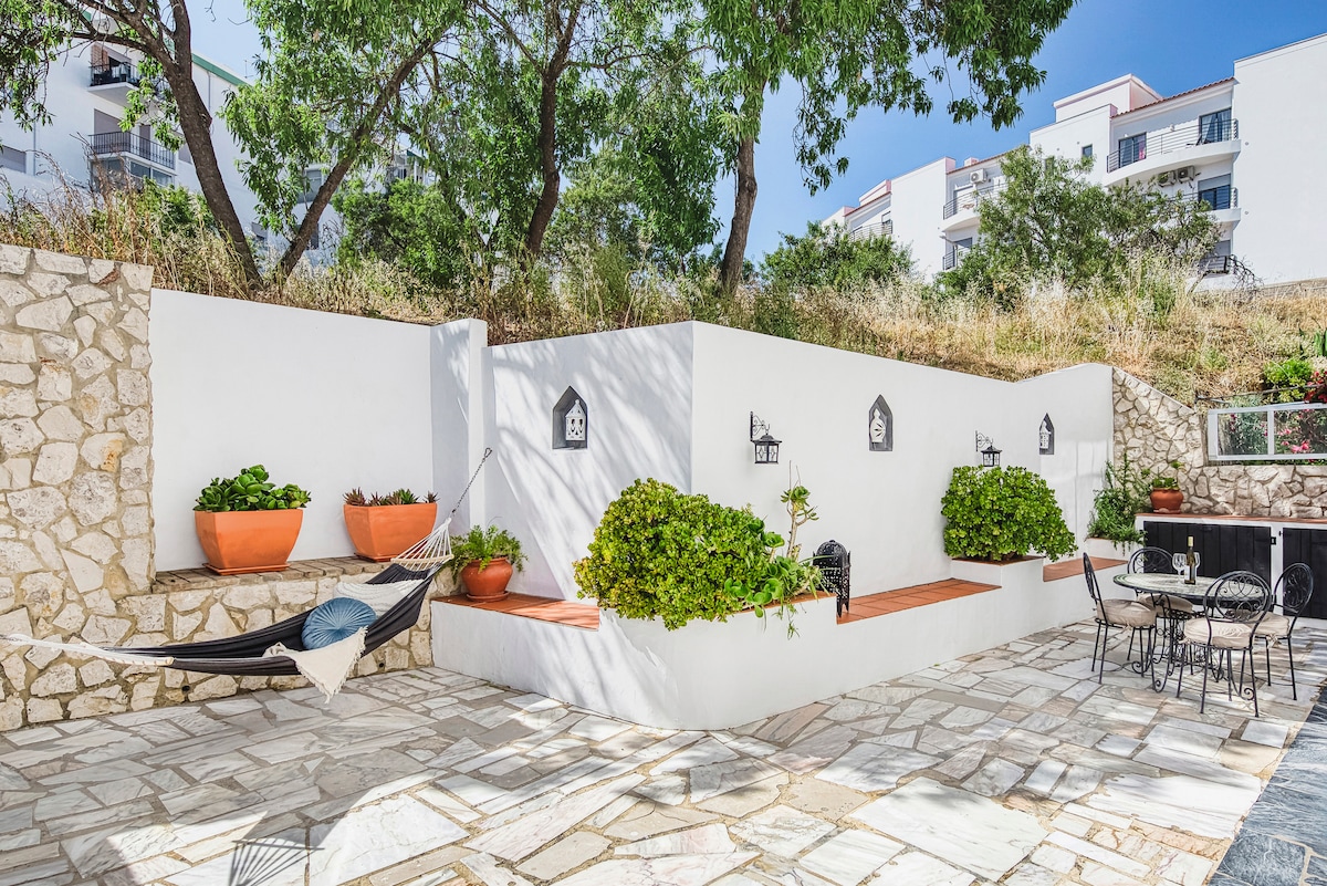 A private terrace is surrounded by greenery, featuring a hammock and seating area. Potted plants add a touch of nature to the white walls. A stone path leads to a dining space with a table and chairs, perfect for enjoying the outdoors.