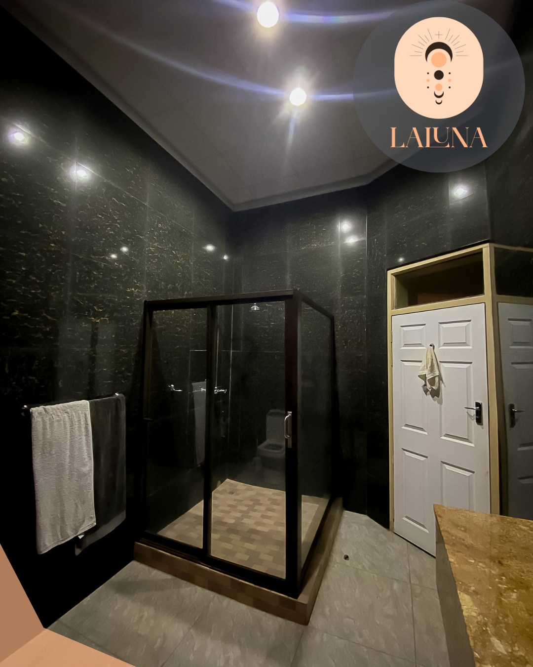 A modern bathroom features a spacious glass shower enclosed by black frames. The walls are adorned with dark tiles, complemented by a light-colored countertop. A white towel hangs on a rack, and a white door leads to additional storage.