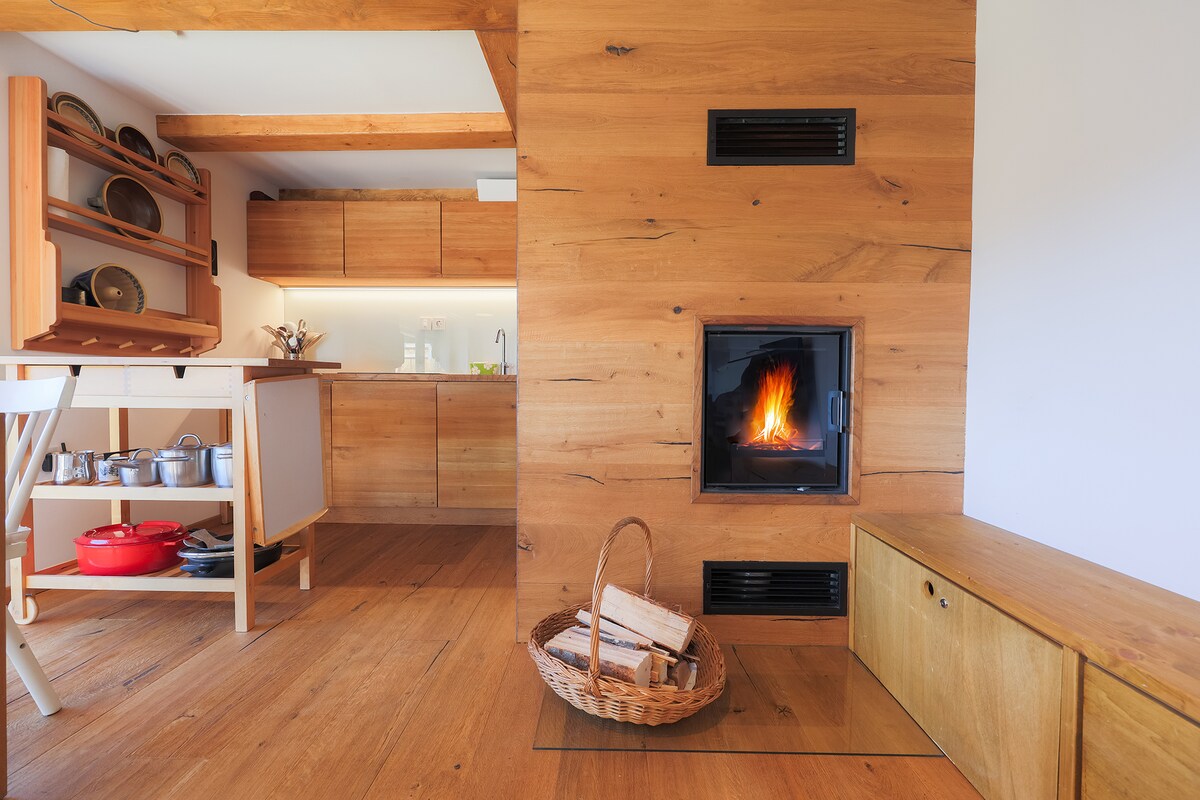 A modern kitchen area features wood elements, with a visible induction cooktop and organized cookware displayed on shelves. A fireplace with a warm flame is centered within a wooden structure, complemented by a basket of firewood nearby. Natural light illuminates the space.