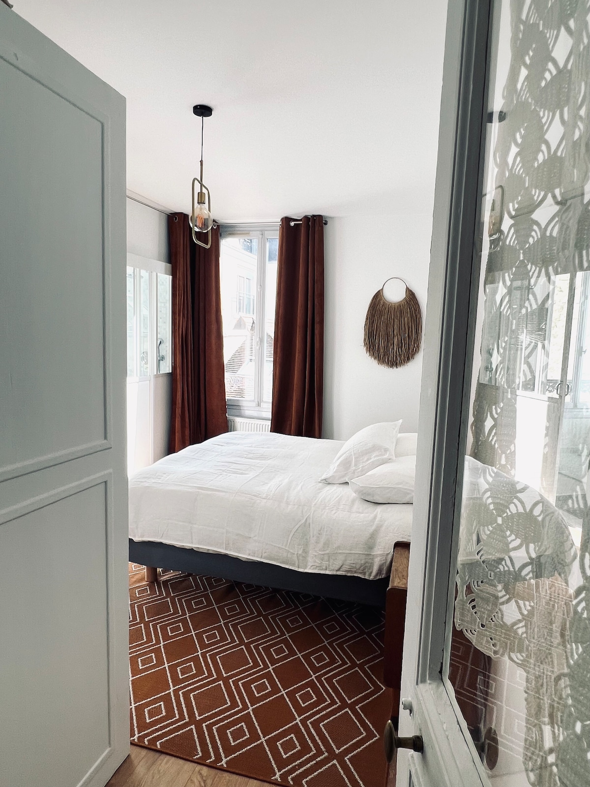 A bright bedroom is accessed through a doorway, featuring a spacious bed dressed in white linens. Earth-toned curtains frame the windows, and a circular wall mirror adds a decorative element. A geometric patterned rug lies beneath the bed, contributing to the room's cozy ambiance.