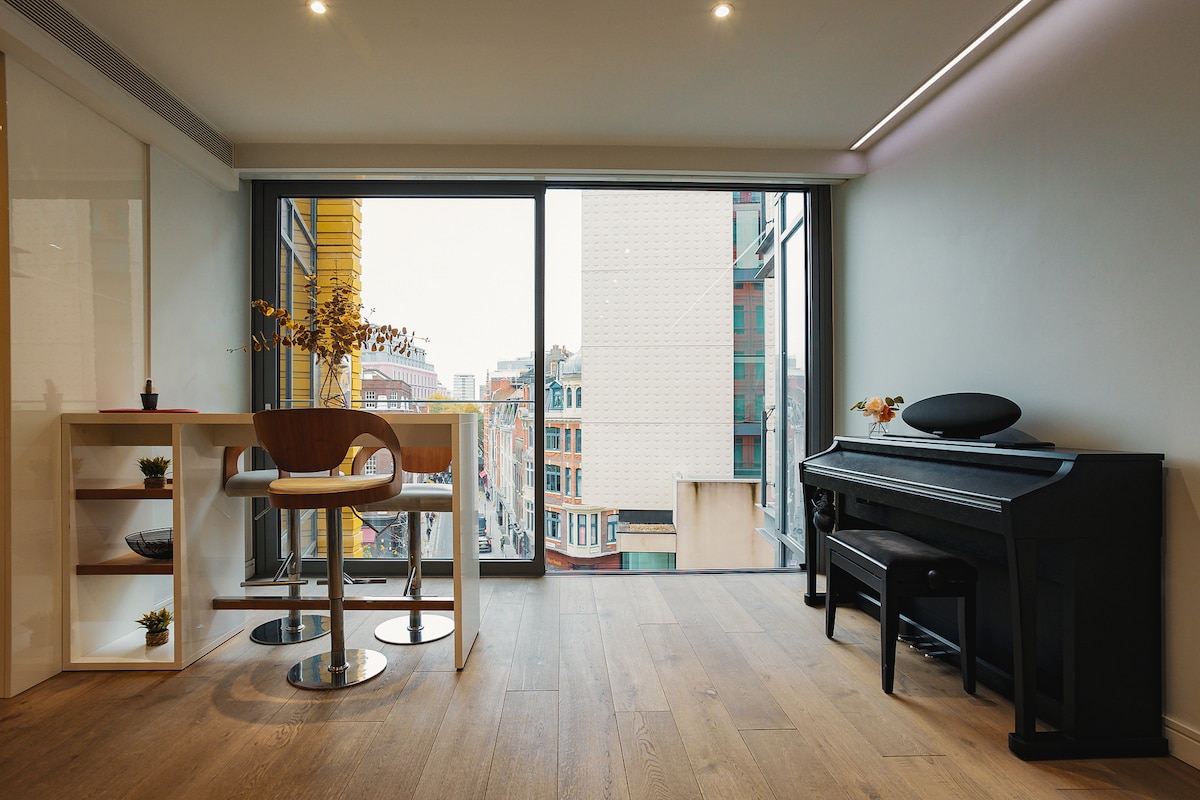 A bright, open space features large windows offering views of the vibrant street below. A simple round table with one chair is positioned near the window, while a sleek black piano is set against the wall, complementing the modern decor.