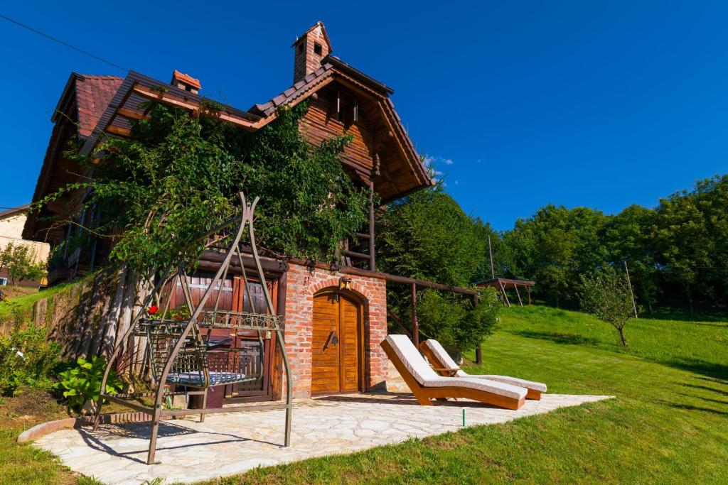A charming wooden holiday home with a brick facade is surrounded by lush greenery. A stone patio features two loungers, inviting relaxation under clear blue skies. A swing set adds a playful touch to the garden, complementing the peaceful atmosphere.