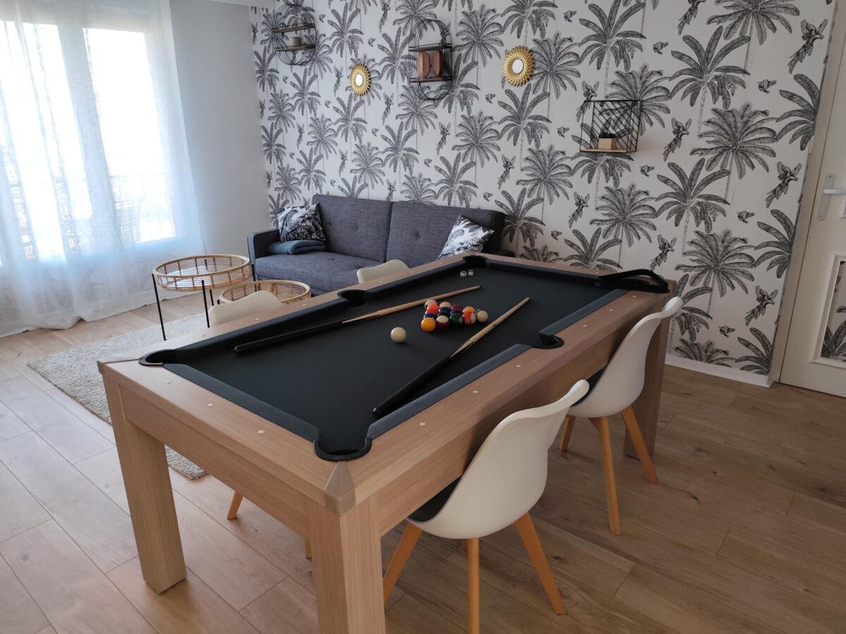 A billiard table is centrally located, surrounded by sleek chairs. An inviting gray sofa sits against a tropical-themed wallpaper. Light filters through floor-to-ceiling windows, enhancing the spacious feel of the living area.