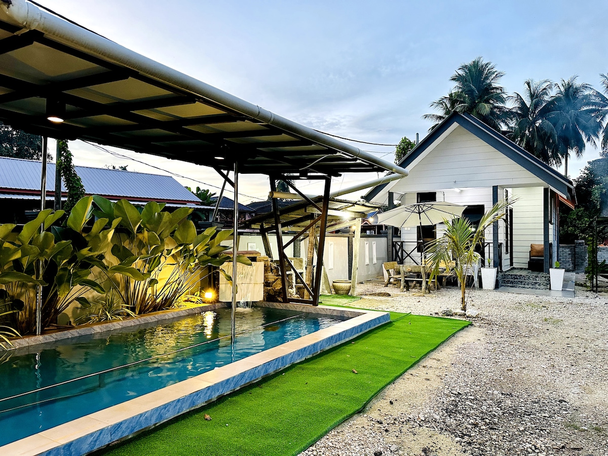 A modern tiny house is surrounded by lush greenery and a well-maintained pool area. The pool reflects evening light, while a covered seating area and umbrella provide shaded outdoor space. The path leads to the entrance, enhancing the welcoming ambiance of the home.