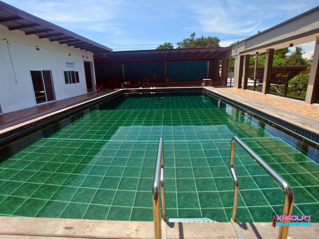 A spacious multi-depth swimming pool is observed, featuring green tiled flooring. Sturdy railings are positioned at the pool's edge for safety. Ample deck space surrounds the pool, providing room for relaxation and sunbathing under a clear blue sky.