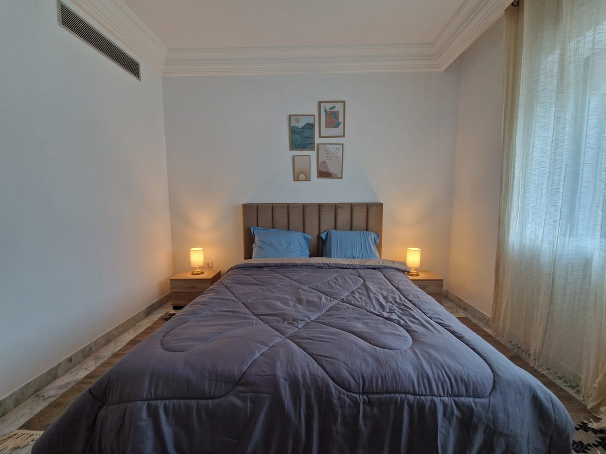 A well-lit bedroom features a spacious bed with a soft blue comforter and matching pillows. Warm light is provided by bedside lamps on either side. Art pieces are displayed above the bed, and gentle drapes frame the window, allowing natural light to filter in.