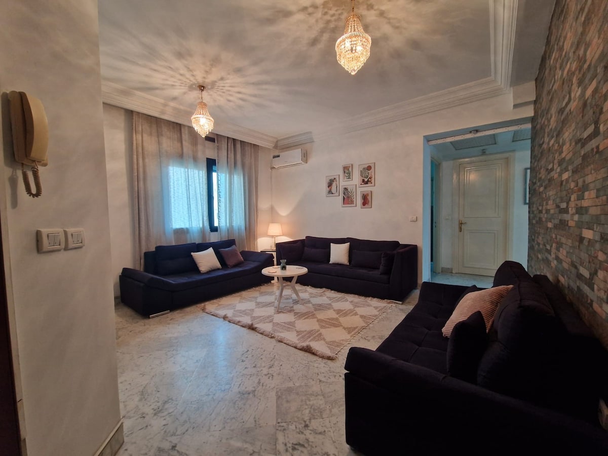 A spacious living area features two dark sectionals arranged around a light circular coffee table. Soft light is diffused through sheer curtains, illuminating a textured stone accent wall. Decorative artwork is displayed on the walls, adding subtle character to the inviting space.