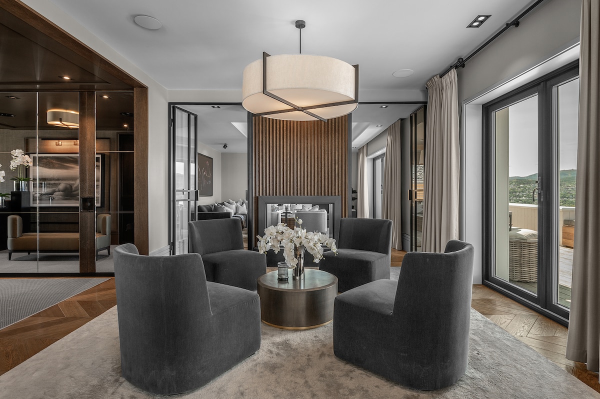 A modern living area is depicted, featuring four plush chairs arranged around a circular table. A large, pendant light hangs from the ceiling, providing illumination. Floor-to-ceiling glass doors lead to a terrace, allowing natural light to fill the space.