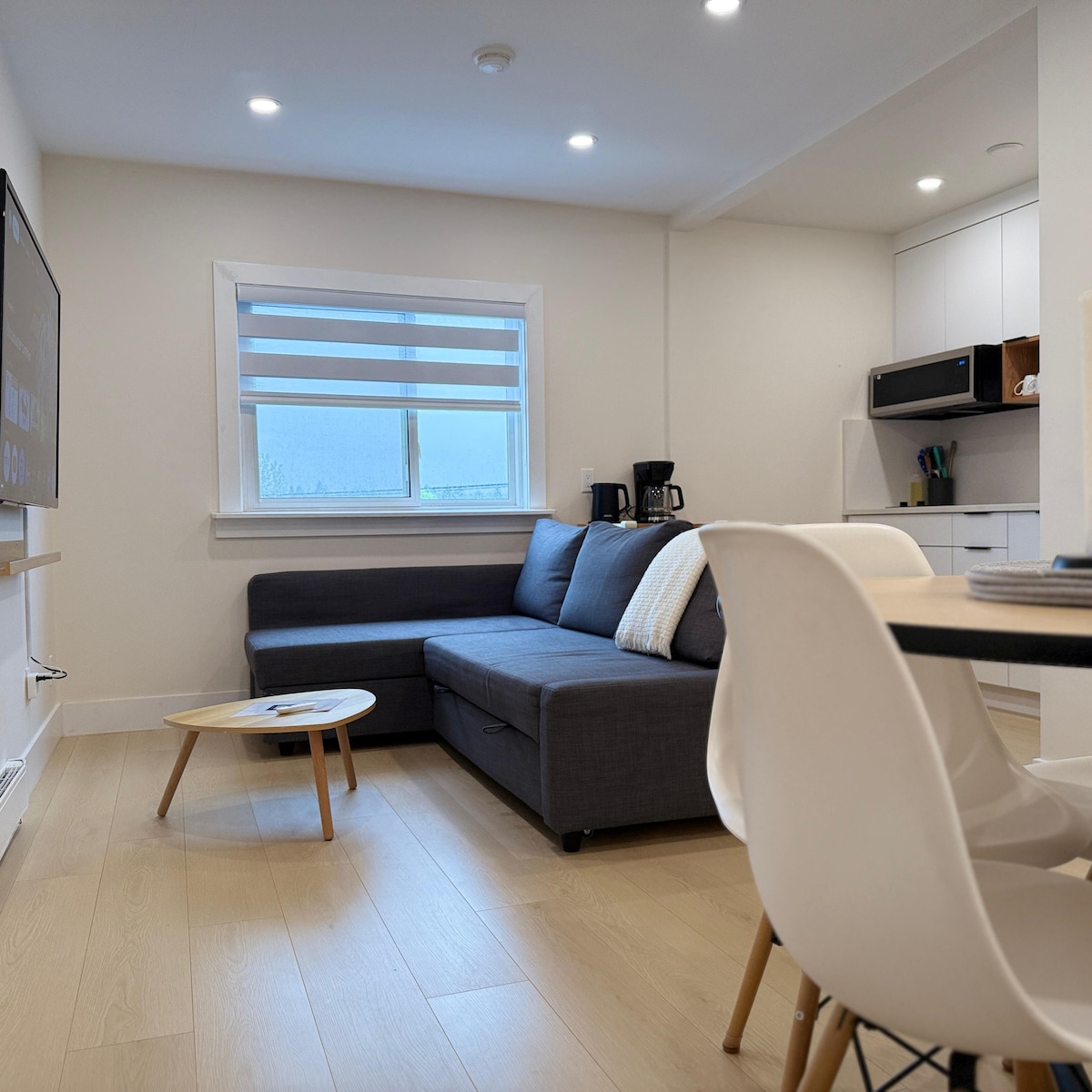 A combined living and dining area is displayed, featuring a dark grey sectional sofa with a light throw pillow. Natural light enters through the window with blinds. A small coffee table sits in front of the sofa, and a kitchenette is visible in the background.