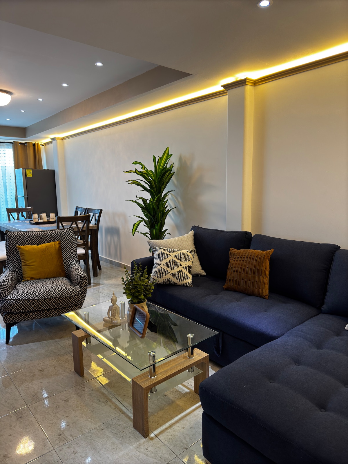 A modern living area features a dark blue sectional sofa paired with a patterned armchair. A glass coffee table is centered, showcasing decorative items. Soft lighting highlights the space, which includes a dining table visible in the background, creating a warm and inviting environment.