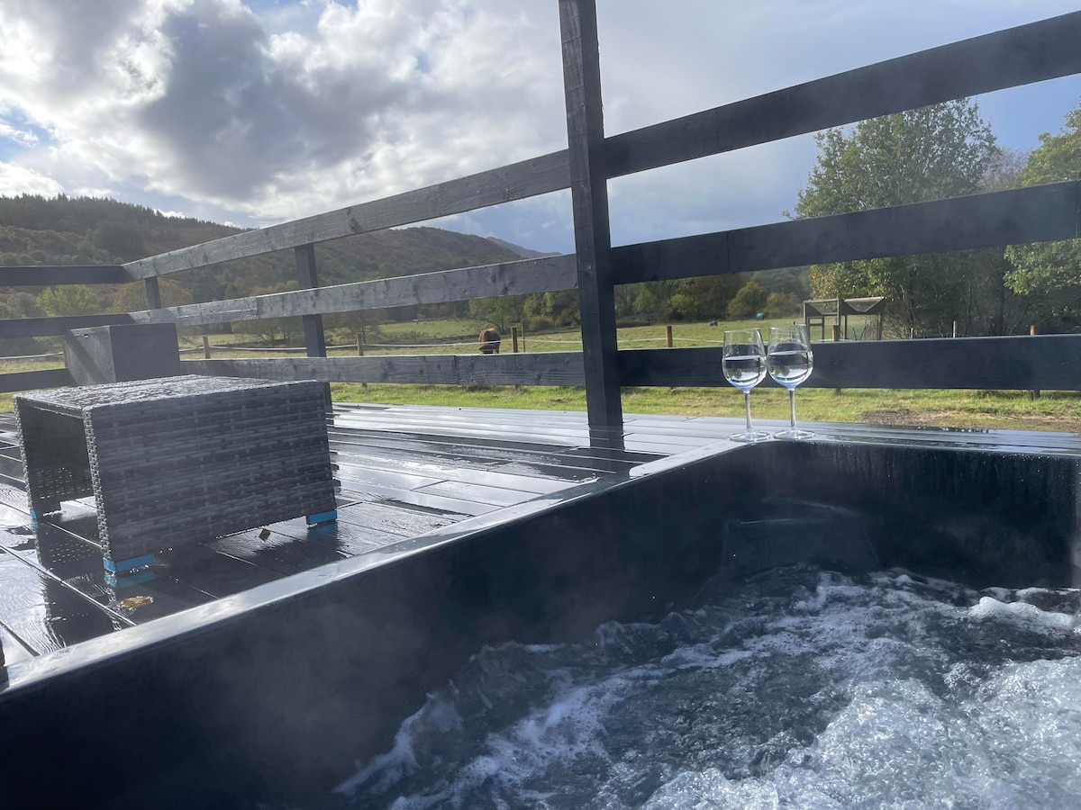 A bubbling hot tub is featured on a wooden deck, with two glasses placed nearby. The backdrop showcases expansive views of rolling hills and greenery, providing a tranquil natural setting.