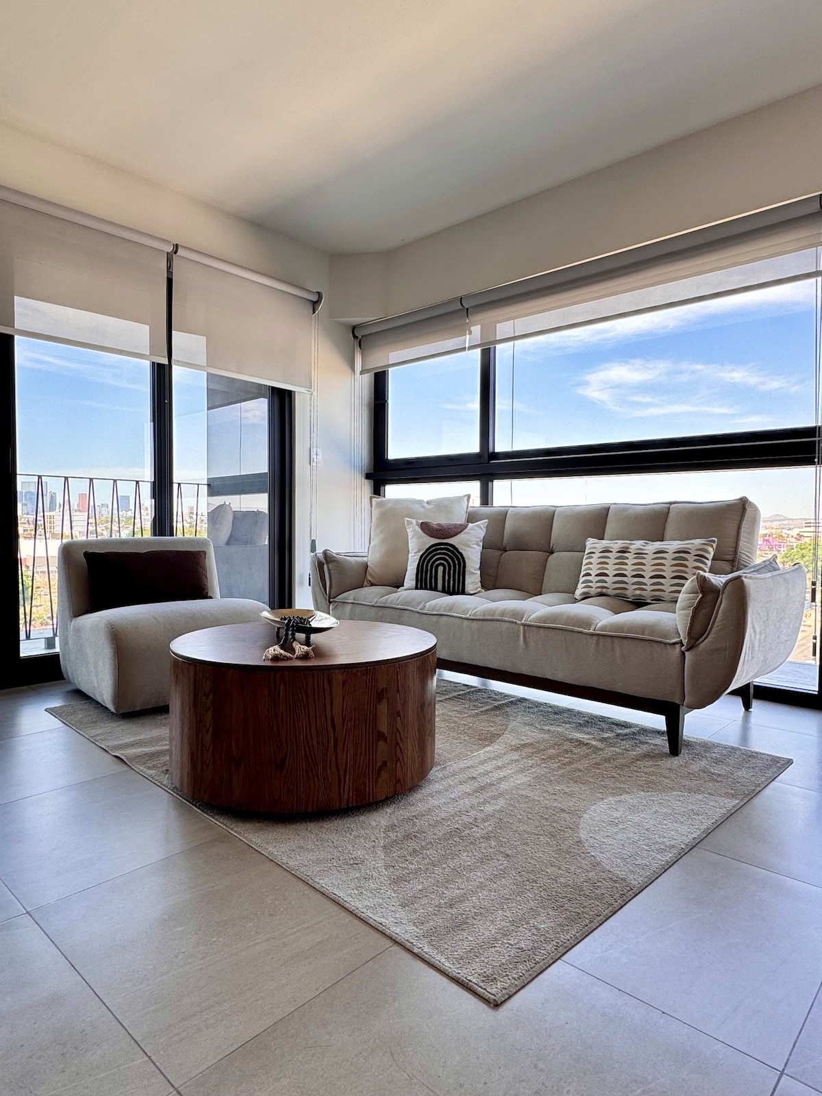 A comfortable seating area features a light-toned sofa and a round wooden coffee table. Large windows allow for ample natural light, while a patterned rug adds texture to the neutral-colored flooring. Scenic views of the surroundings are visible through the glass panels.
