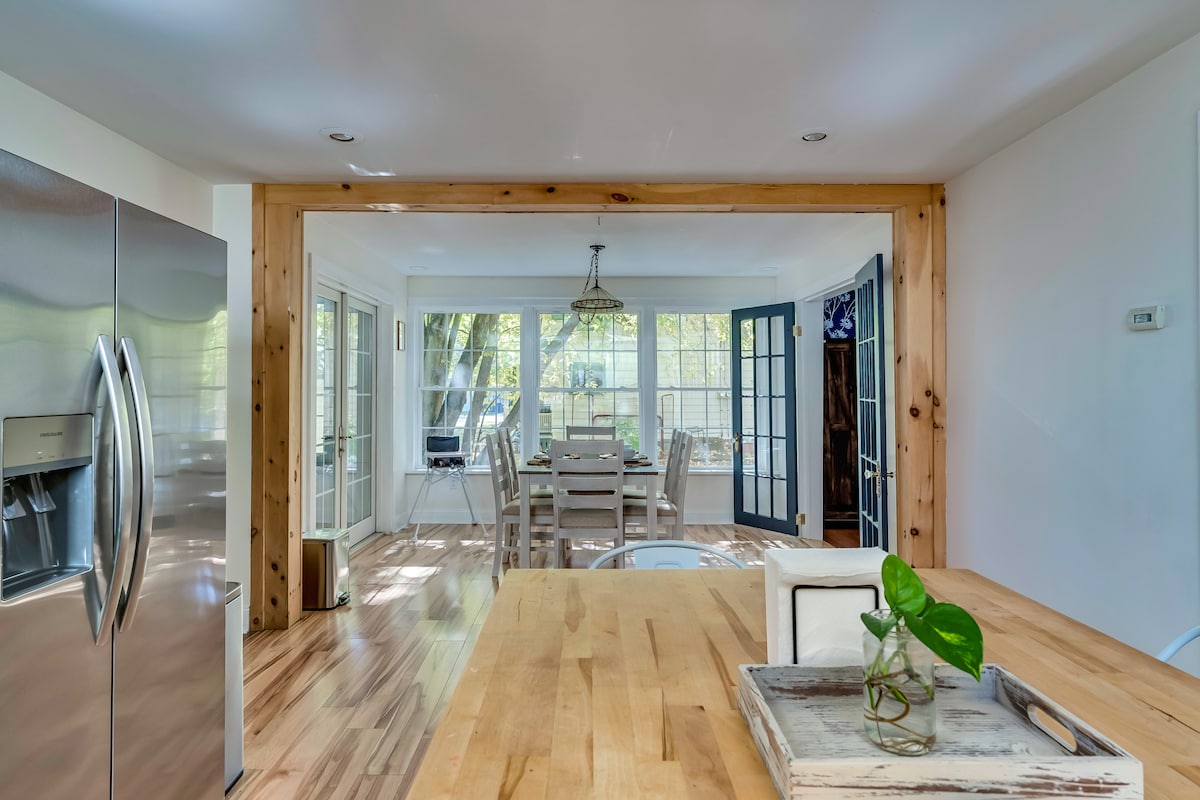 A bright and airy kitchen area features a wooden island with a built-in tray, and stainless steel appliances are visible in the background. Large windows invite natural light, while a dining area with a wooden table and six chairs can be seen through an open doorway.