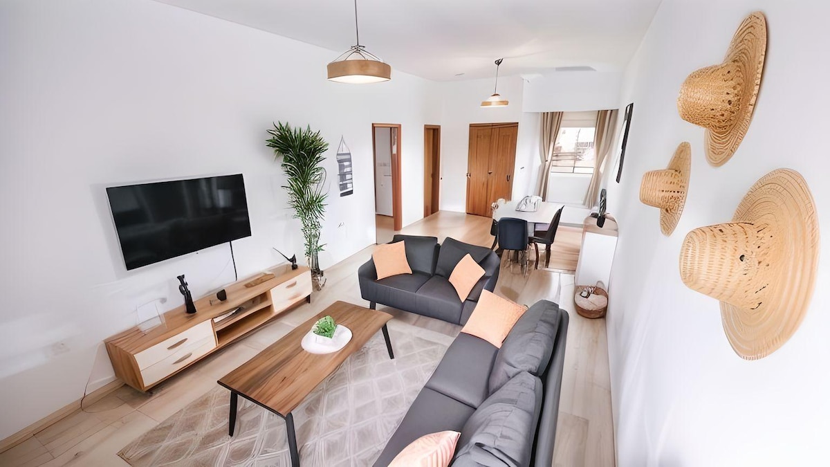 The living area presents a spacious layout with a comfortable grey sofa, accented by orange cushions. A wooden coffee table is centered on a patterned rug. A large television is mounted on the wall, and decorative hats are displayed beside the windows, which provide natural light.