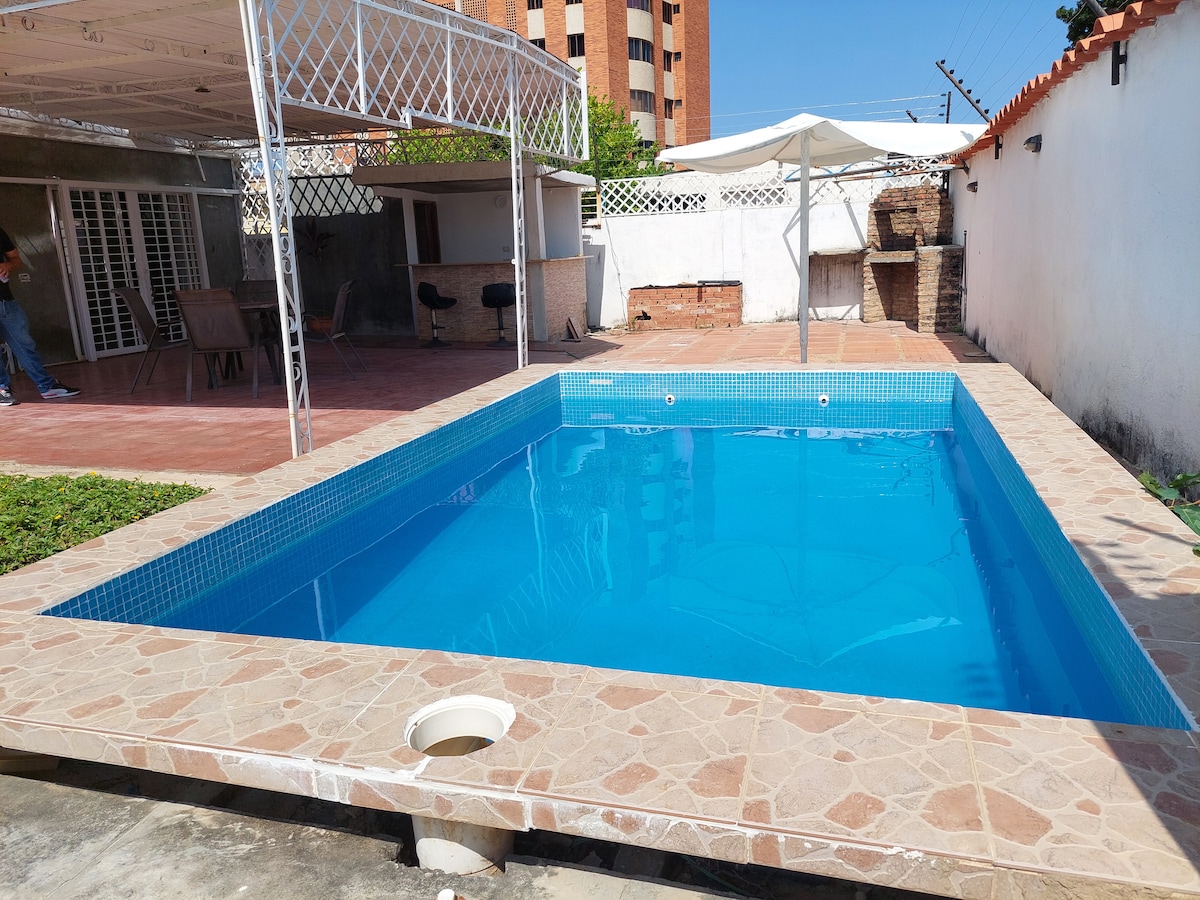 A refreshing swimming pool is set within a private outdoor area, featuring clear blue water surrounded by tiled decking. A shaded seating area can be seen in the background, along with a patio that provides space for relaxation.