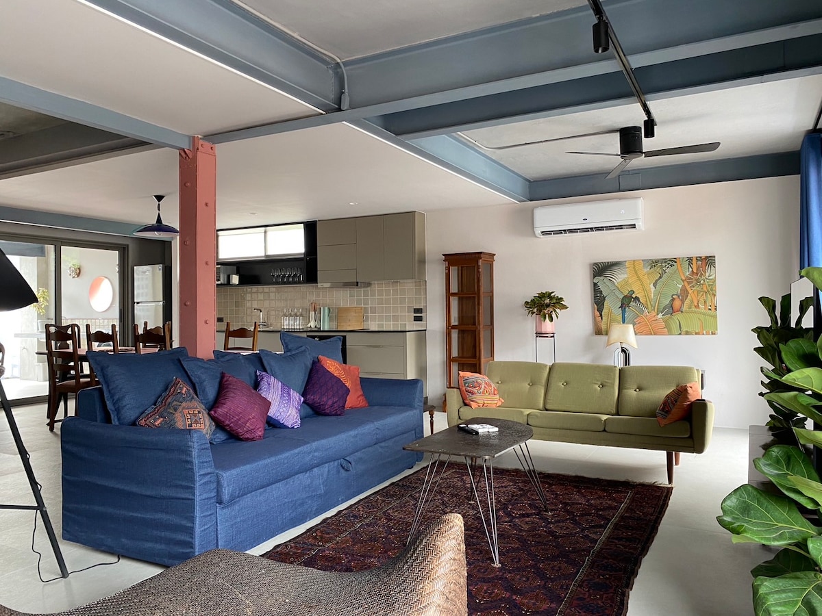 A spacious living area showcases a blue sofa adorned with decorative pillows, alongside a green couch. A coffee table sits atop a patterned rug, with a fan overhead. The kitchen is visible in the background, featuring modern cabinetry and a dining table.