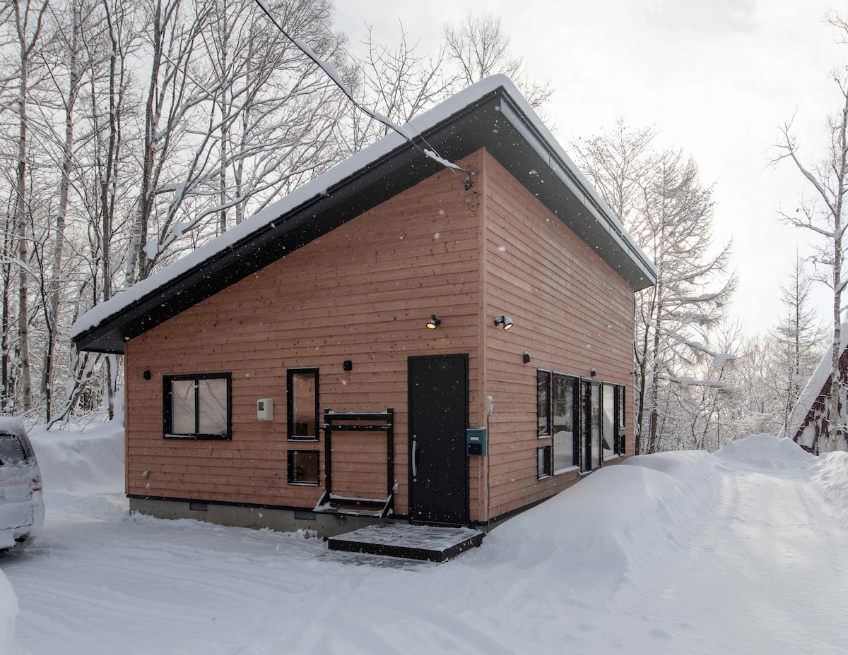 A modern cottage is nestled in a snowy landscape, featuring a wooden exterior with large windows. Snow gently falls on the surrounding area, and a driveway is visible, leading to a parked vehicle. The roof is sloped, contributing to the structure's contemporary design.