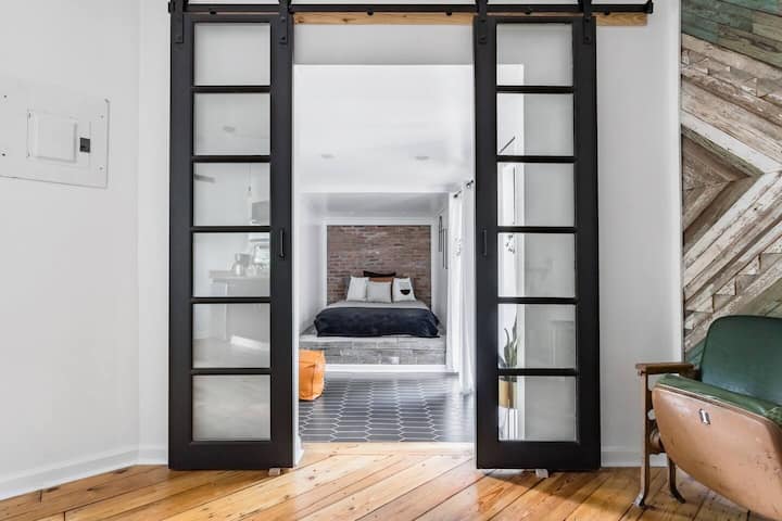 Štýlová vstupná hala s elegantnými čiernymi posuvnými sklenenými dverami, ktoré sa otvárajú do svetlej spálne. Priestor má teplé drevené podlahy, geometrické dlaždice a útulnú posteľ orámovanú stenou s textúrou.