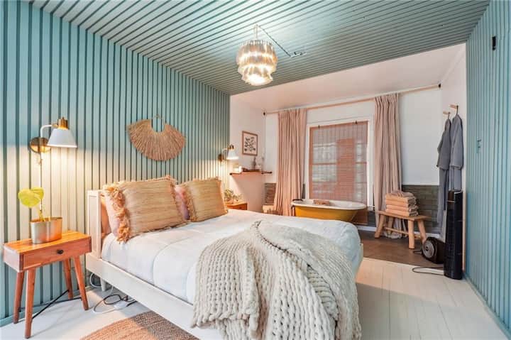 Štýlová spálňa s modernou, ale útulnou atmosférou s odvážnym dizajnom obložených panelov, ktoré siahajú od steny až po strop. V izbe nájdete teplý drevený nočný stolík, tkané dekorácie, mäkkú neutrálnu posteľnú bielizeň a očarujúcu voľne stojacu vaňu v pozadí.