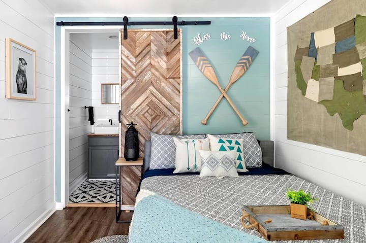Útulná spálňa s modernými rustikálnymi dizajnovými prvkami s drevenými stodolovými dverami, stenami s lodnými doskami a hravými dekoratívnymi prvkami. Jemné modré a neutrálne tóny vytvárajú pokojné a príjemné útočisko.