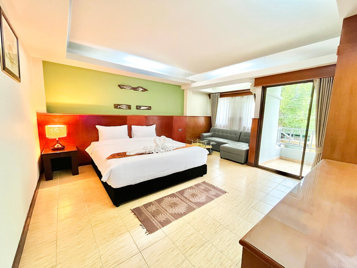 A spacious guest room is presented with a large bed covered in white linens, complemented by subtle decorative accents on the walls. A seating area features a gray sofa, and large windows allow natural light to fill the space. A coffee table and decorative rug add warmth to the ambiance.