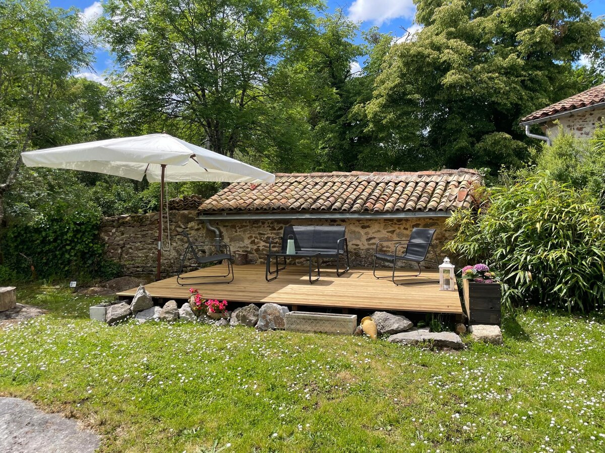 A tranquil outdoor terrace features a wooden deck adorned with a lounge set and a large umbrella for shade. Surrounding the area, lush greenery and colorful flowers complement the setting, with a traditional stone wall and tiled roof in the background.
