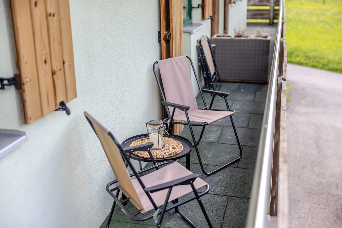 A peaceful outdoor seating area is featured, with two folding chairs positioned around a round table. The table is adorned with a glass lantern, and the setting is surrounded by a well-maintained, grassy landscape.