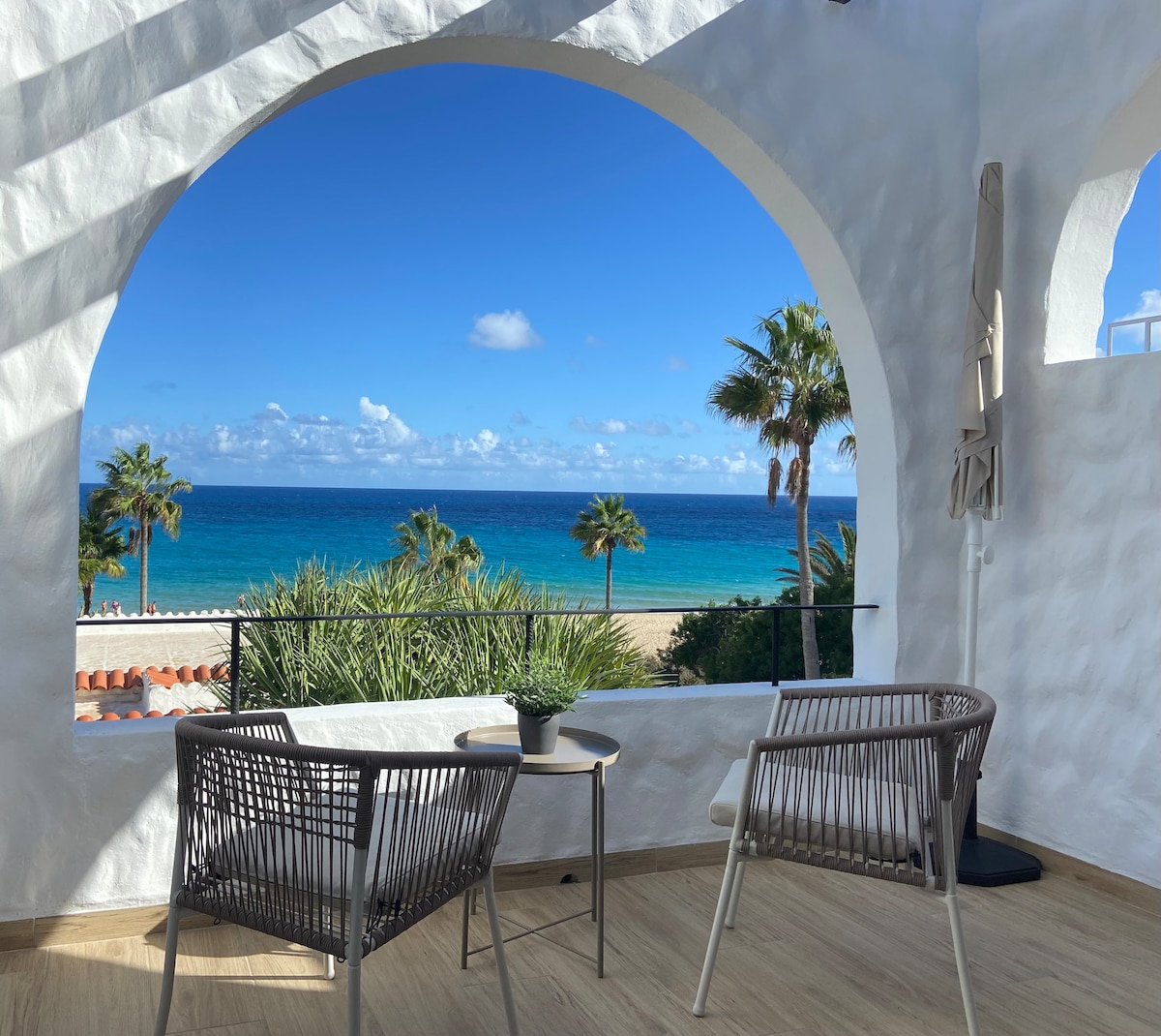A spacious balcony features two woven chairs and a small round table, set against a backdrop of clear blue skies and the expansive sea. Palm trees frame the view, enhancing the connection with the coastal landscape.