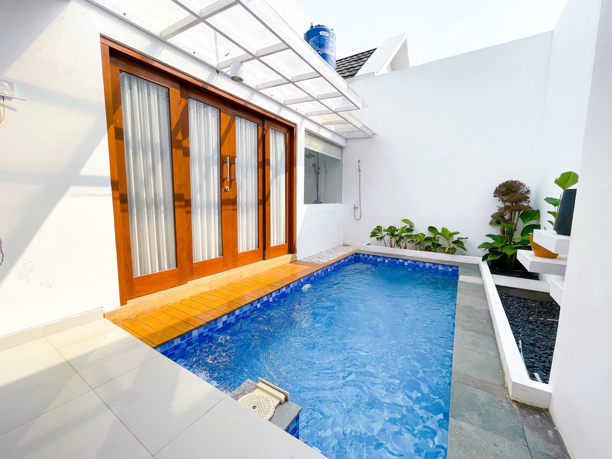A private enclosed swimming pool is surrounded by minimal landscaping, featuring green plants. The pool area is decorated with wooden sliding doors that lead to a bright interior, and natural light reflects off the water's surface.