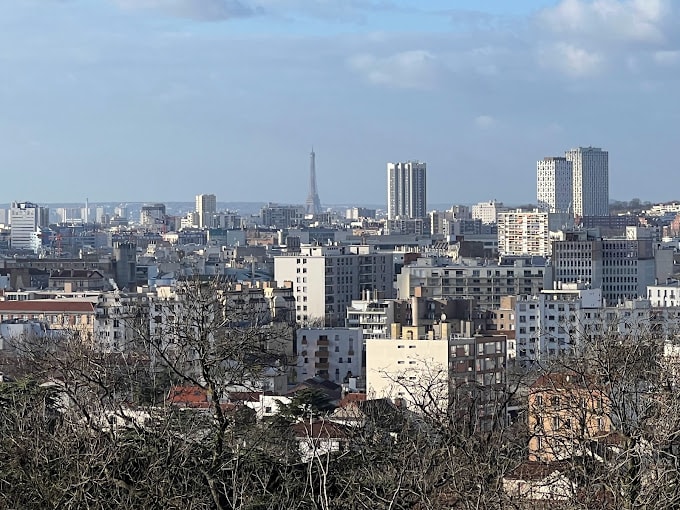 A panoramic view of a city skyline is captured, showcasing various residential and commercial buildings. The Eiffel Tower is visible in the distance, surrounded by urban architecture. Trees and greenery are seen in the foreground, providing a contrast to the cityscape.