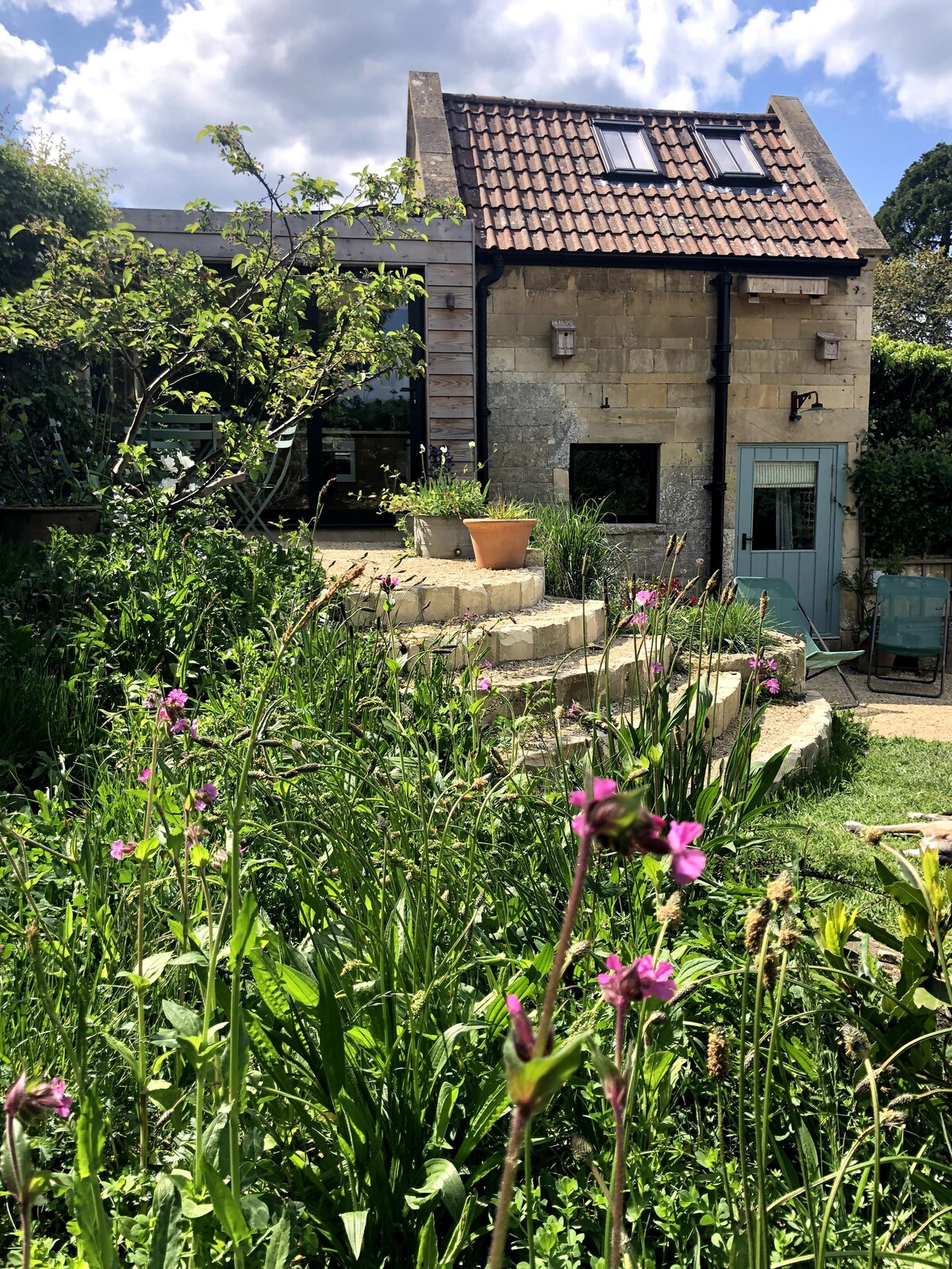 The exterior of the property features a quaint stone building with a slate roof, surrounded by lush greenery and colorful wildflowers. A series of steps leads to the entrance, while bright sky and clouds provide a serene backdrop.