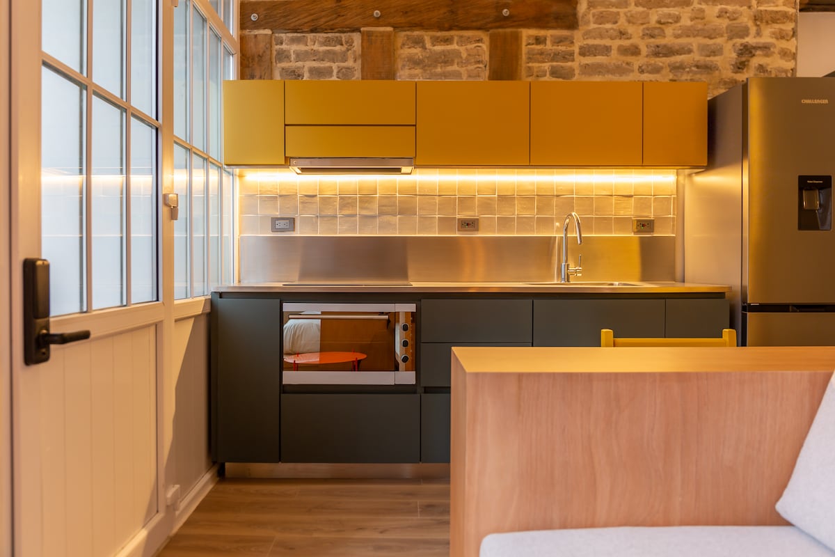 The kitchen features a contemporary design with yellow overhead cabinets complemented by dark lower cabinets. A stainless steel countertop and backsplash are illuminated by warm lighting. Essential appliances, including an oven and refrigerator, are included. Stone walls add texture to the modern space.