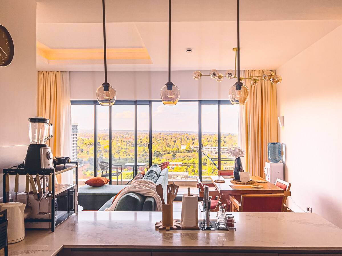 A modern kitchen with a sleek marble countertop features a blender and various utensils. Behind, a spacious living area is highlighted by tall windows offering expansive views of greenery. Soft drapes frame the windows, and stylish lighting fixtures add a contemporary touch.