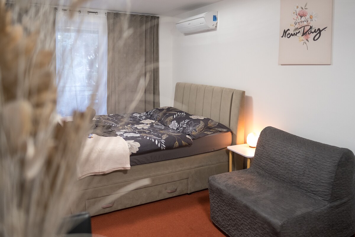 A spacious sleeping area features a large bed with floral bedding, complemented by a cozy chair in the corner. A soft-colored blanket is draped over the bed. Natural light filters through sheer curtains, creating a gentle ambiance in the room.