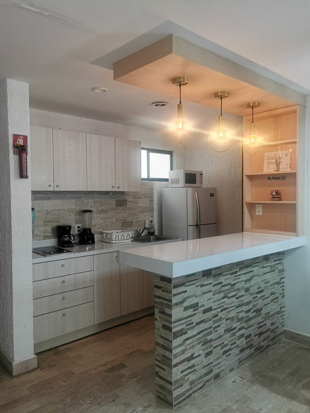 A modern kitchen is equipped with light wood cabinetry and a sleek countertop. Pendant lights hang above the breakfast bar, providing illumination. Appliances include a refrigerator and a blender, while natural light enters through a window, enhancing the open feel of the space.