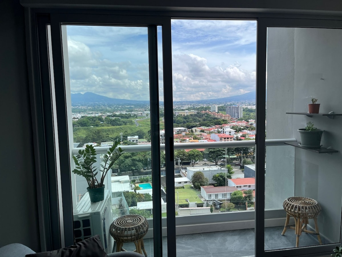 A large glass door opens to a balcony with a view of rolling green hills and the city skyline. Potted plants are visible on the railing, while cozy seating is positioned nearby. The vibrant landscape includes houses, a swimming pool, and distant mountains.
