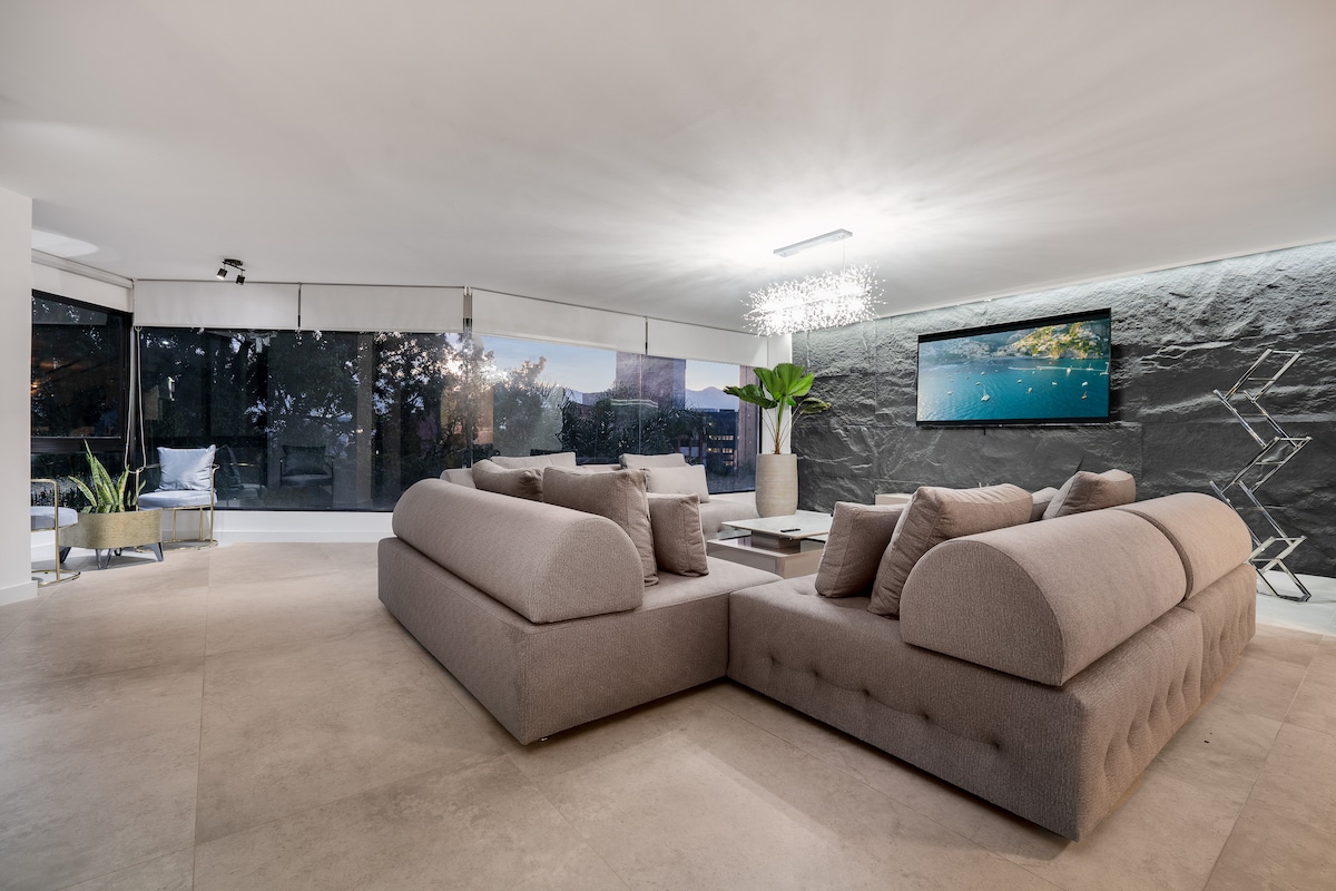 A spacious living area features a large, light-colored sectional sofa with rounded edges. A flat-screen television is mounted on the wall, while natural light floods in through expansive windows adorned with light shades. Decorative plants add a touch of greenery to the space.