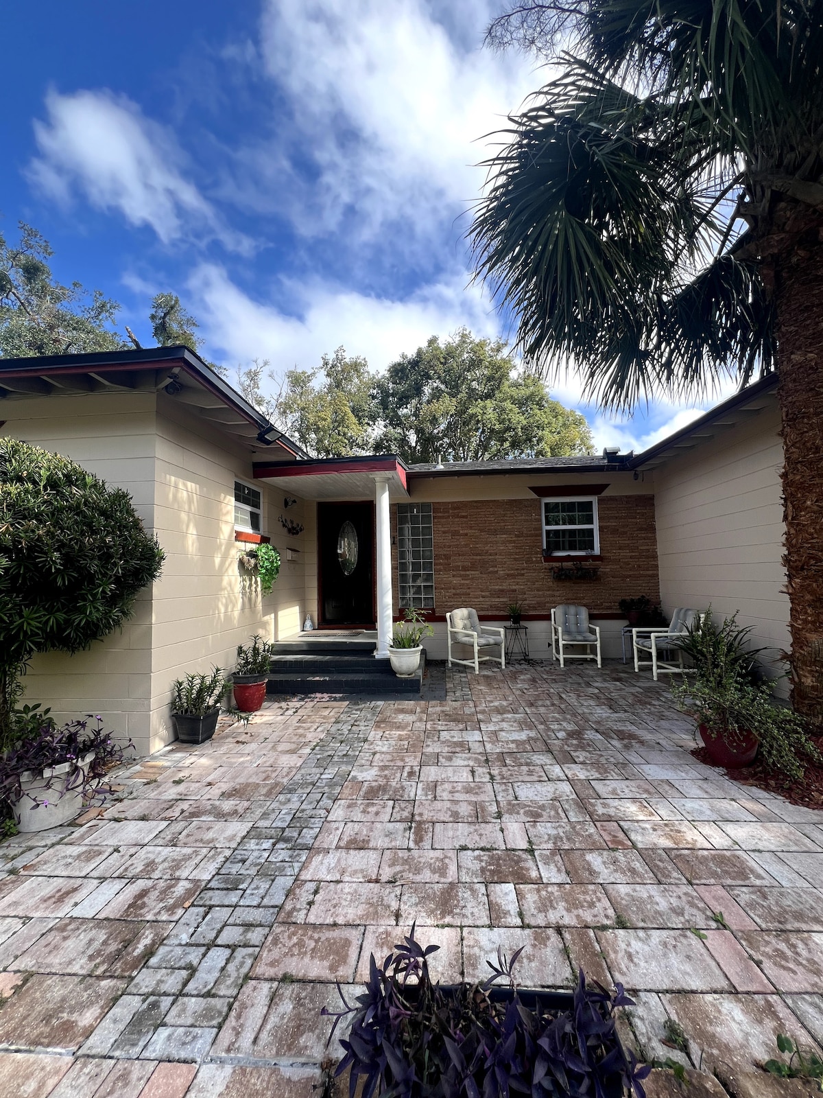 A welcoming front yard is presented, featuring a patterned stone pathway leading to a covered entrance. Potted plants are arranged thoughtfully around the space, with white chairs positioned for relaxation under the shade of palm trees and a clear blue sky above.