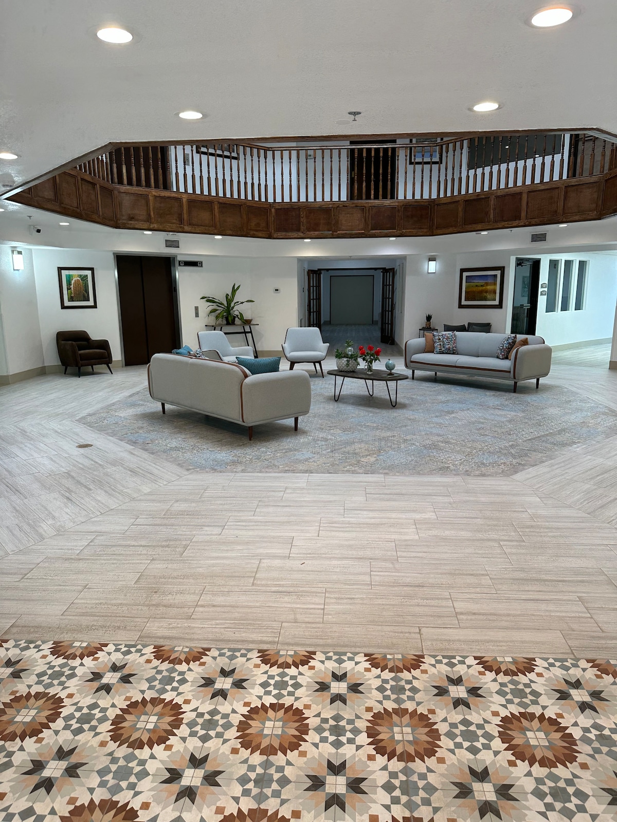 An open lobby space showcases modern furnishings, including soft seating arrangements on a patterned area rug. A decorative table with fresh flowers is positioned in the center, while natural light floods the room through large windows. The unique tile work adds visual interest to the floor.