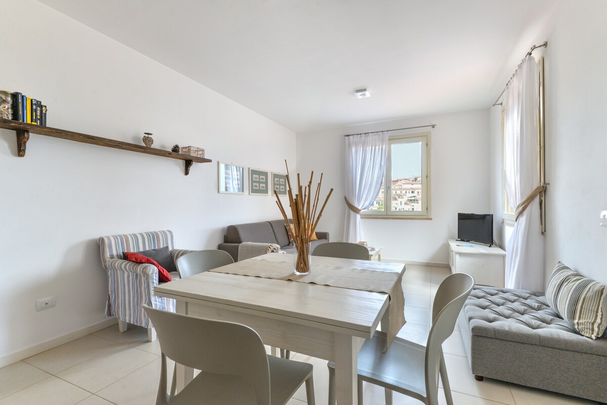 A bright living area features a spacious dining table surrounded by light chairs. A comfortable gray sofa and armchair are present, along with a television on a stand. Natural light fills the room through a window, and decorative items are arranged on a wall shelf.