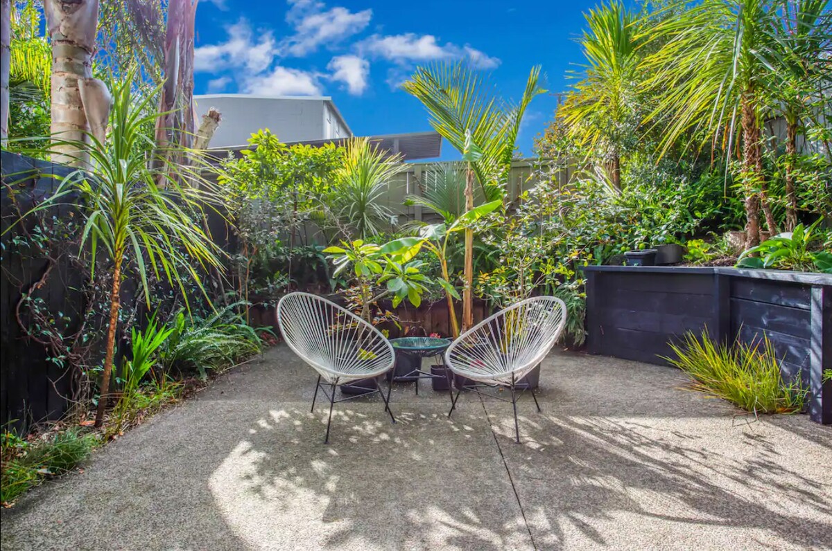 A peaceful courtyard garden is showcased, featuring two modern chairs surrounded by lush greenery and tropical plants. Natural sunlight filters through the leaves, creating soft shadows on the textured ground. The area offers a serene setting for relaxation or social gatherings.